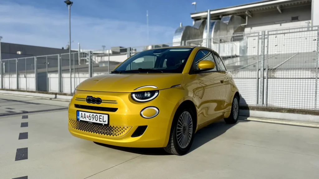 Žltý Fiat 500 Hybrid stojí na slnečnom betónovom parkovisku pred kovovým plotom a budovou, nasnímaný spredu zľava s viditeľnou tabuľkou EÚ.